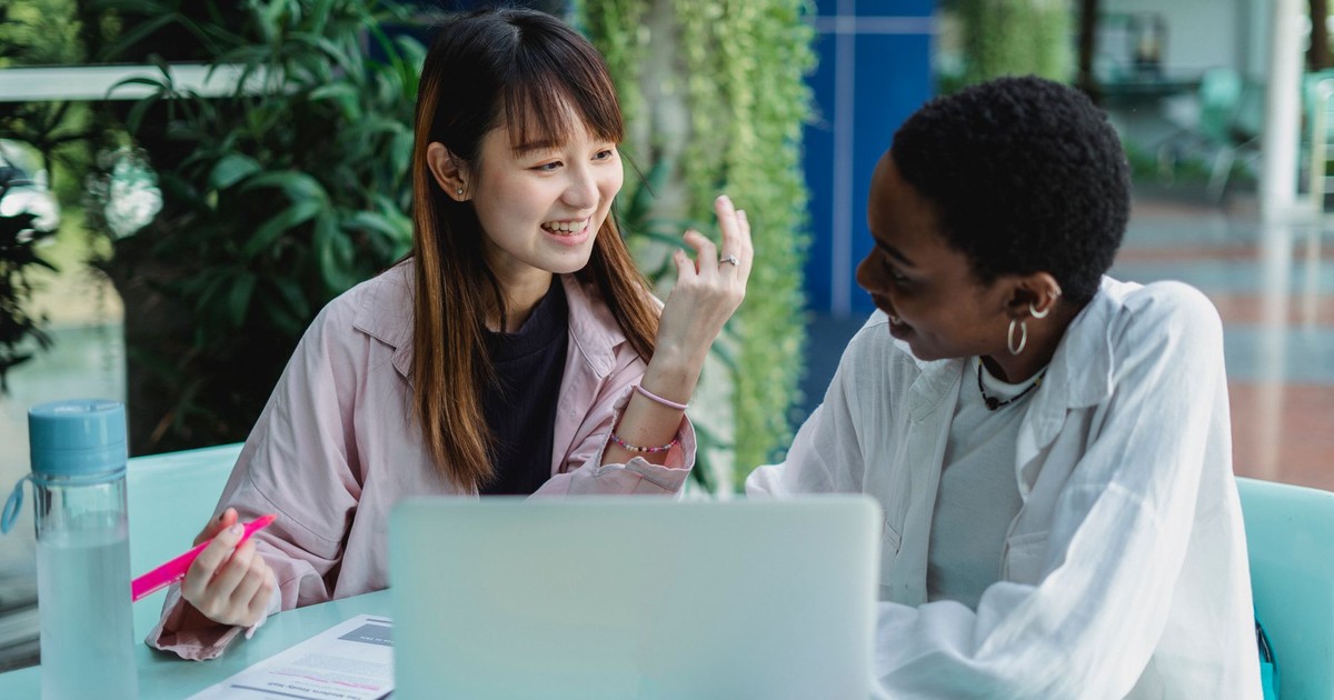 Young glad multiracial girlfriends talking while doing homework at table with netbook and looking at each other in garden