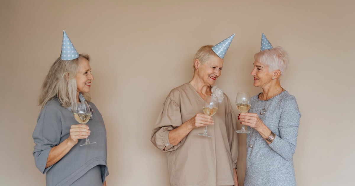 Three senior women in party hats celebrating with drinks indoors.