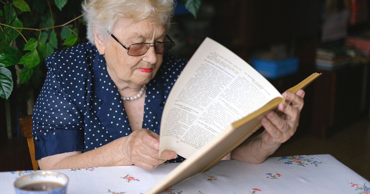 Serious female pensioner in eyeglasses reading book while sitting at table with cup of hot drink in room on blurred background