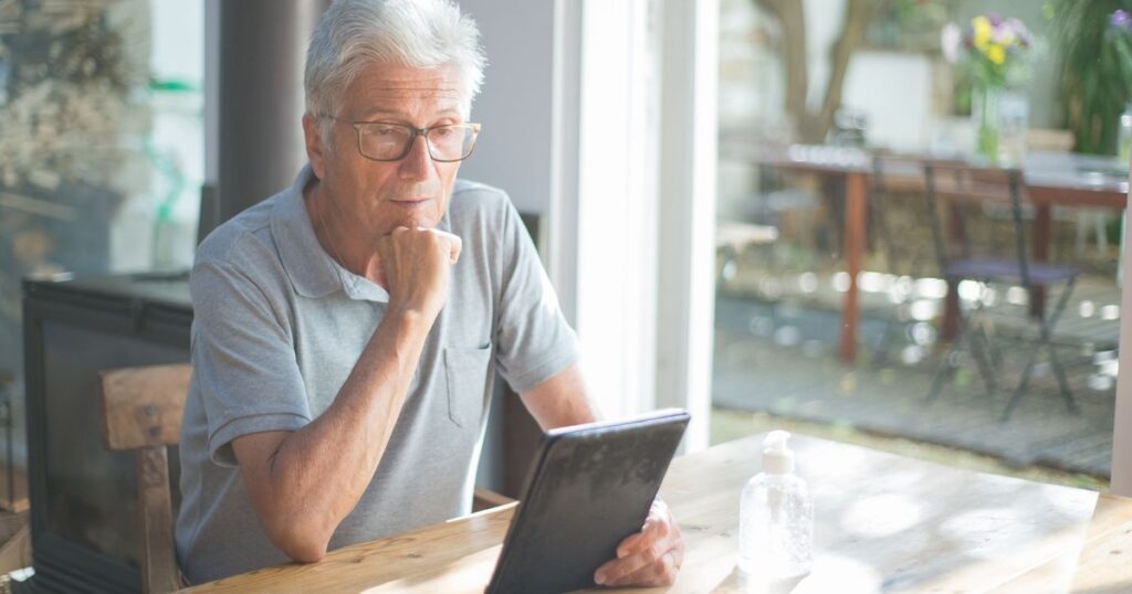 Senior man with eyeglasses reading a tablet at home in a sunlit room.
