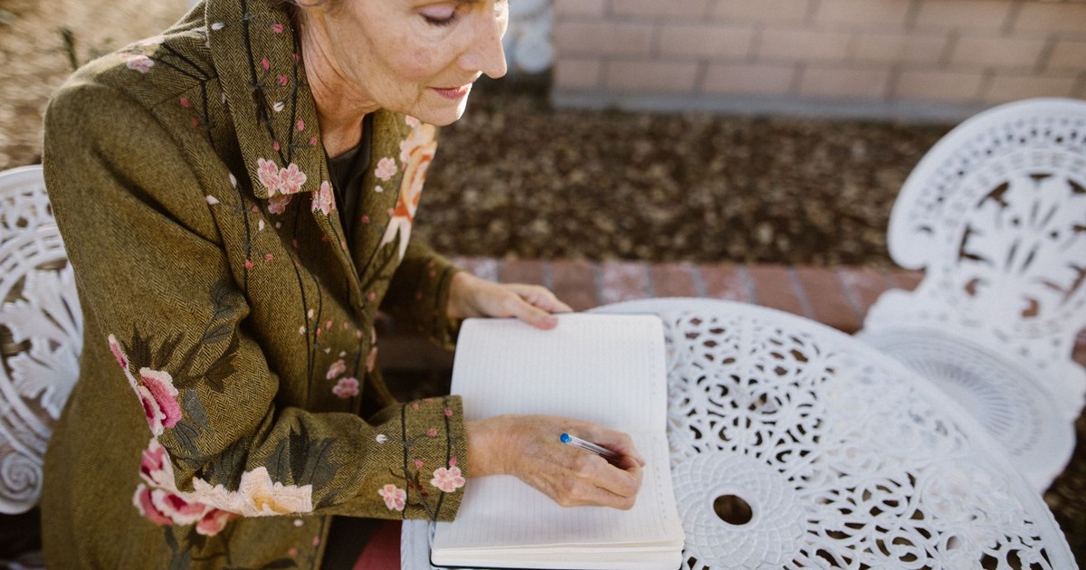 Senior woman sitting outdoors writing in a notebook, enjoying a peaceful garden setting.