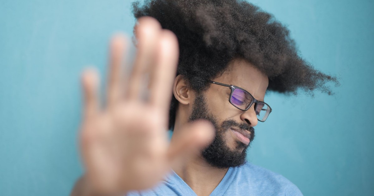 Man with afro hair and glasses making a stop gesture against blue background.
