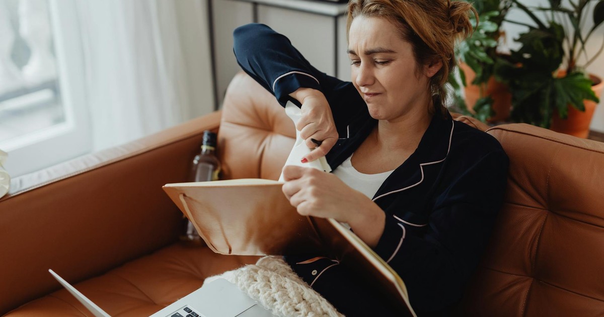 Woman in pajamas tearing paper on a sofa, expressing frustration. Indoors setting with laptop nearby.