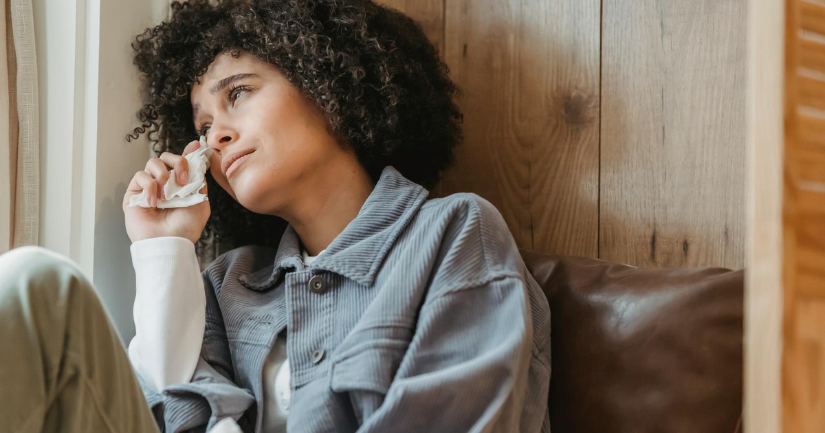 Desperate African American female with curly hair wiping face with tissue while crying from sadness on windowsill and looking away