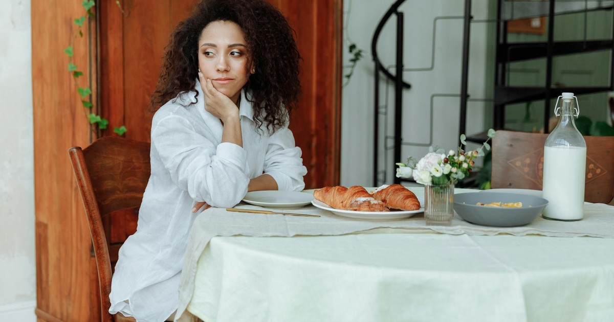 A woman with curly hair sits at a breakfast table with croissants and milk.