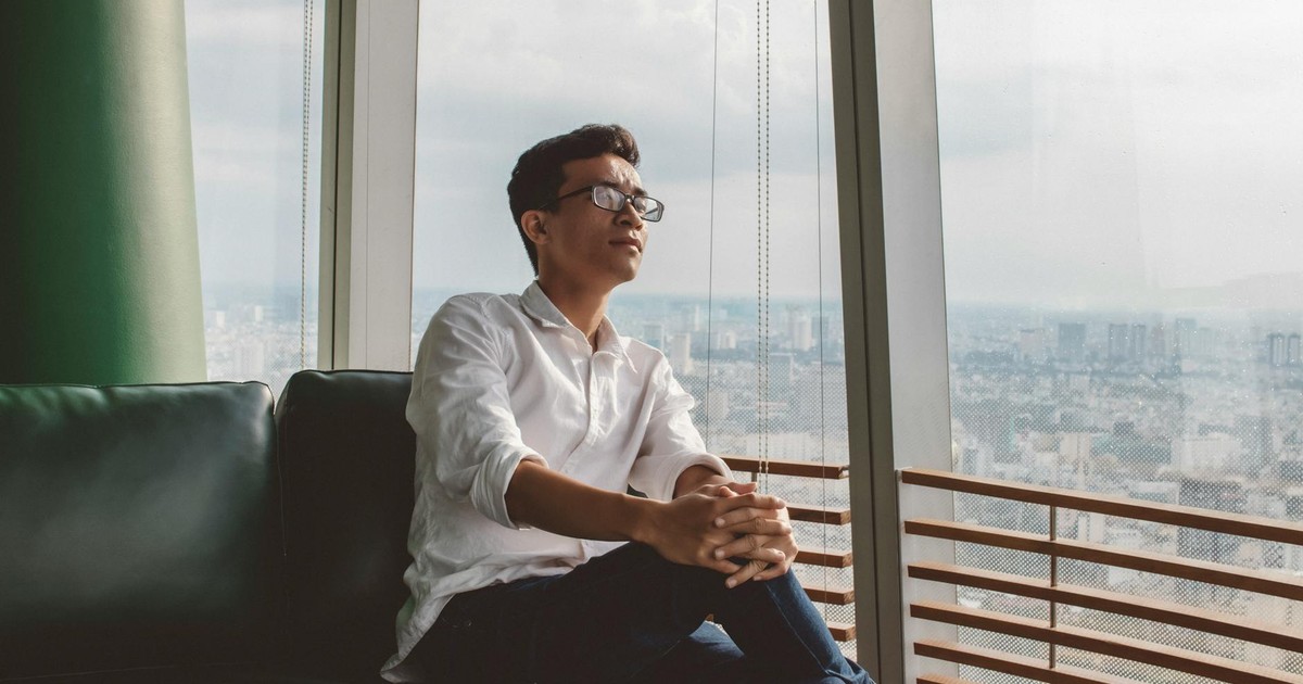 A man in a white shirt sits thoughtfully by a large city view window, enjoying the afternoon light.