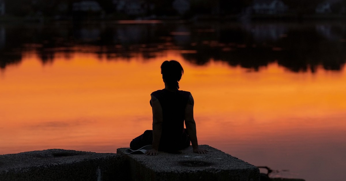 A person meditates at sunrise by a calm lake, creating a serene silhouette.