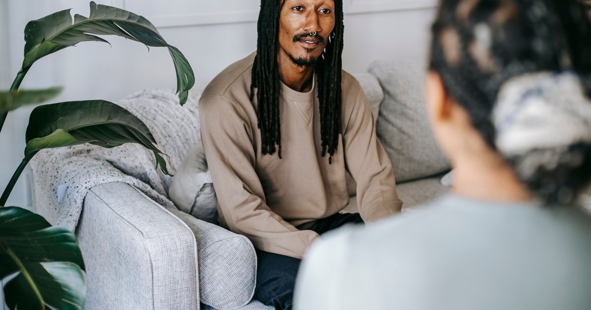 Content African American male with dreadlocks wearing casual outfit having conversation with female friend while sitting on couch in cozy living room