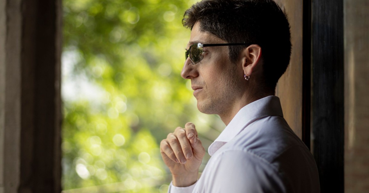 A stylish man wearing sunglasses gazes thoughtfully out a window, framed by greenery.