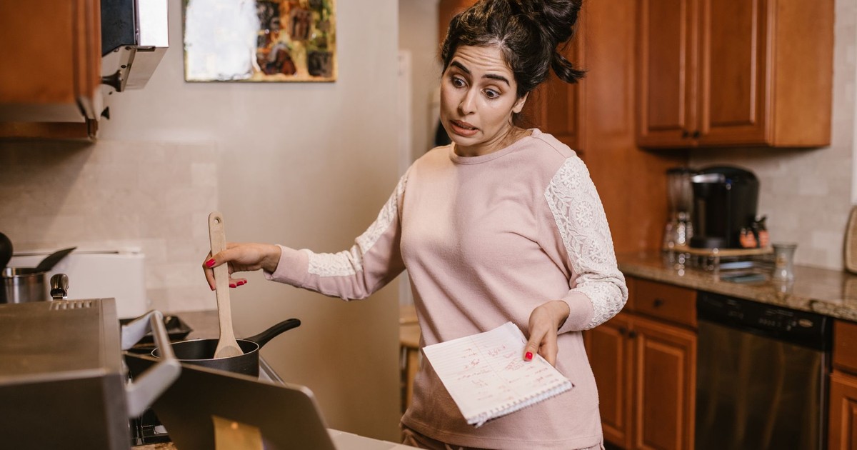 Woman multitasking in her kitchen with a laptop and notebook, cooking and working from home.