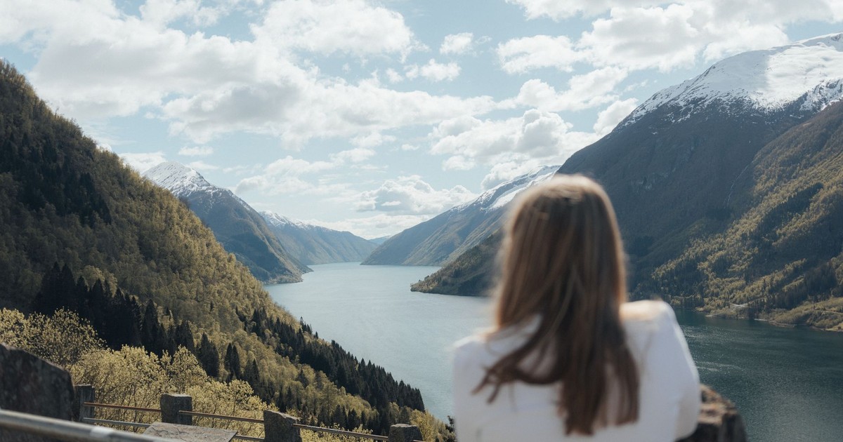 Woman enjoying a panoramic view of a Norwegian fjord surrounded by mountains, under a clear spring sky.