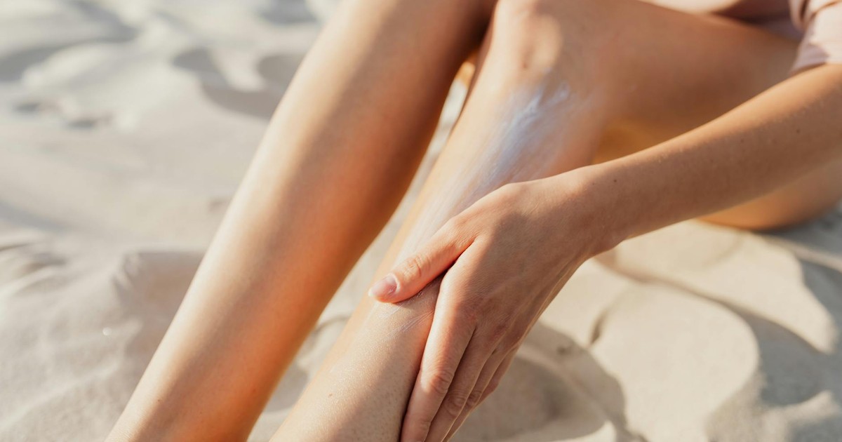 Close-up of a woman applying sunscreen on her legs while sitting on the beach sand.