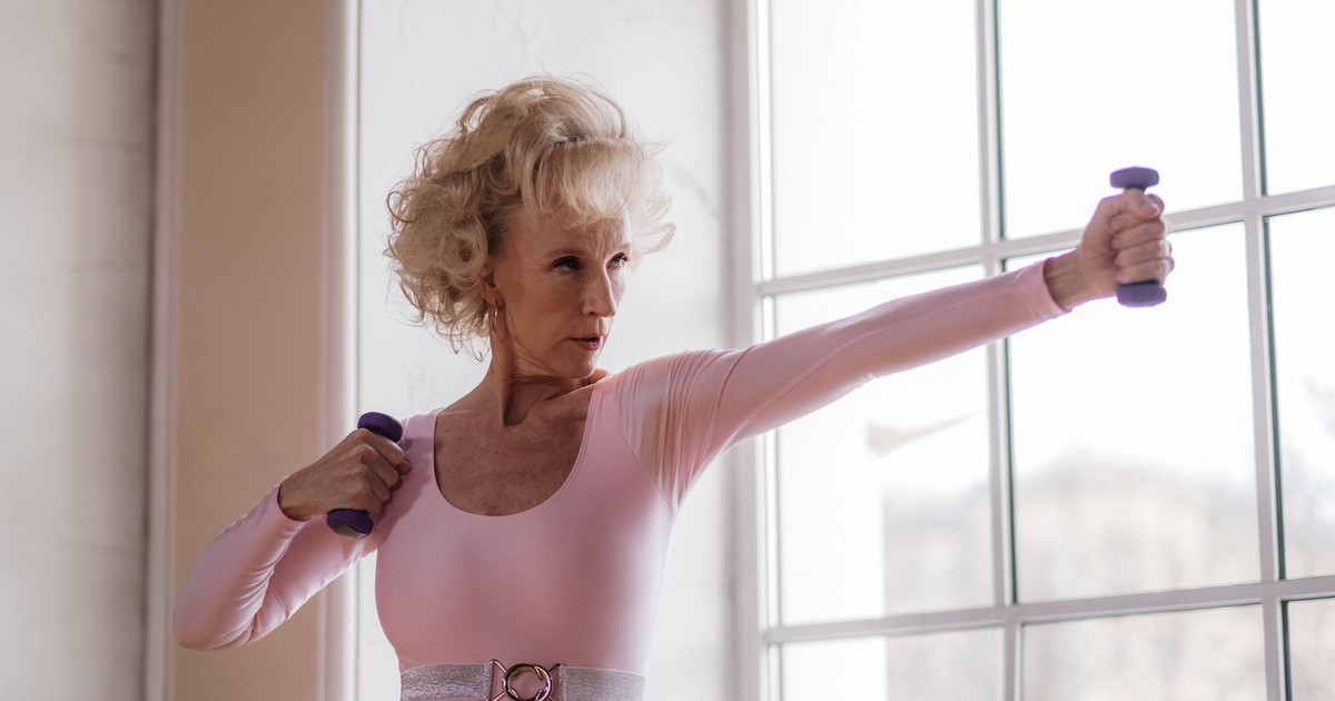 Senior woman in pink leotard working out with dumbbells near a large window indoors.