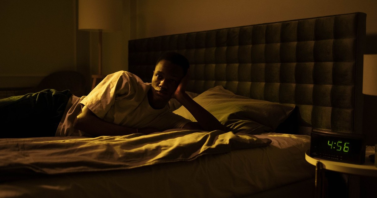 A man lies awake in bed, illuminated by early morning light from a clock reading 4:56 AM.