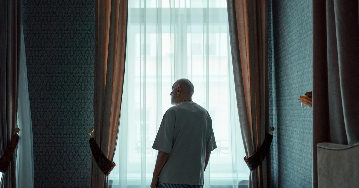 Elderly man standing by a window in an elegant room, looking outside. Ideal for concepts of isolation or contemplation.
