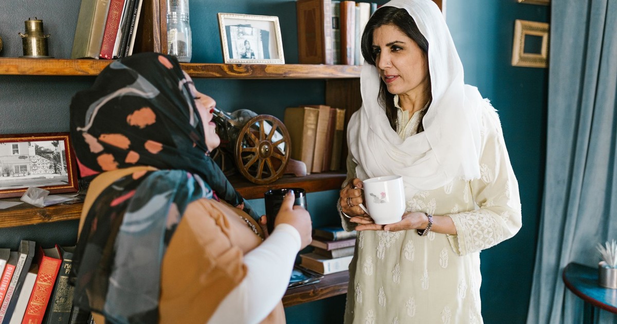 Two women in traditional clothing share a conversation over coffee mugs in a cozy room.