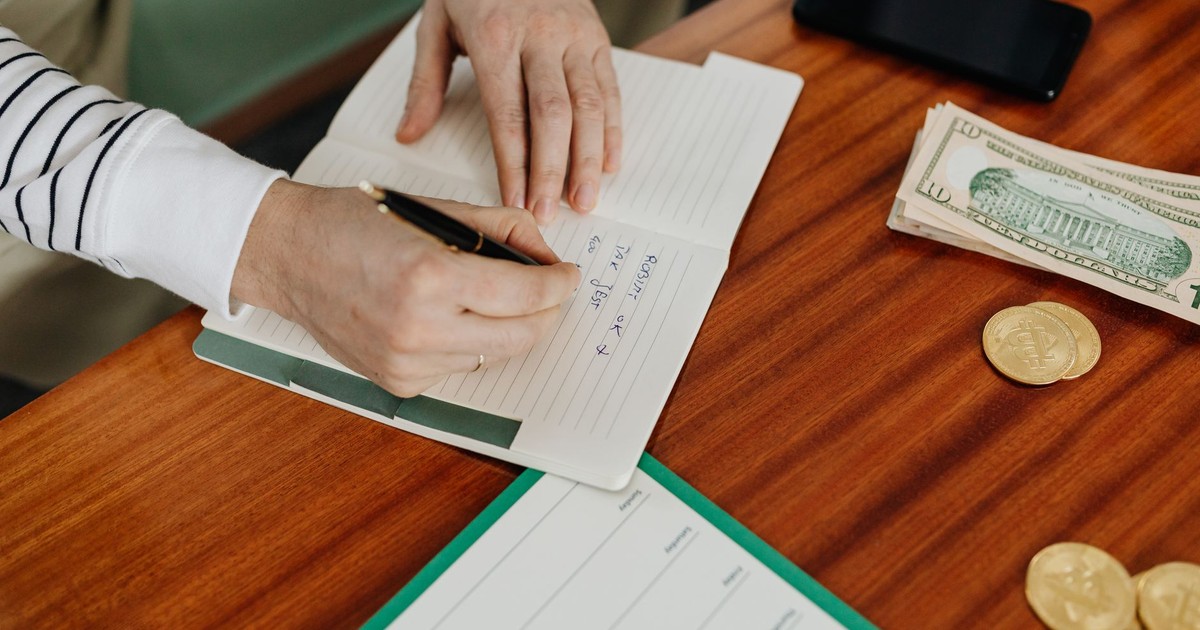 Person writing in a notebook on a wooden table with cash, coins, and a phone nearby.