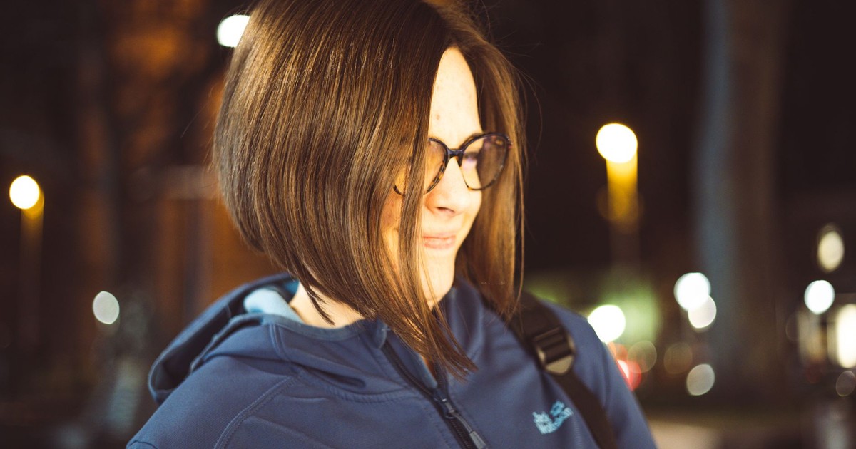 A woman with bob hairstyle and eyeglasses smiles outdoors at night, illuminated by streetlights.