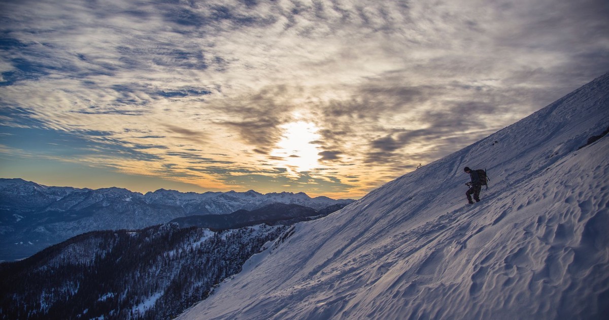 A hiker scales a snowy mountain slope at dawn near Radovljica, Slovenia, capturing a serene winter landscape.