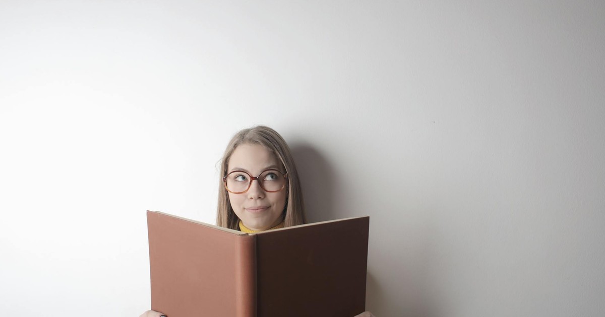 Positive young curious female wearing eyeglasses looking up while standing against gray wall with big opened book with brown cover in hands