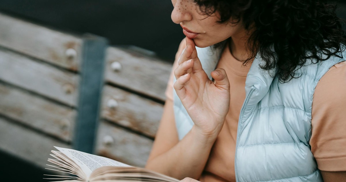 From above of crop young female with dark curly hair in casual clothes touching lips and reading interesting book om bench in park