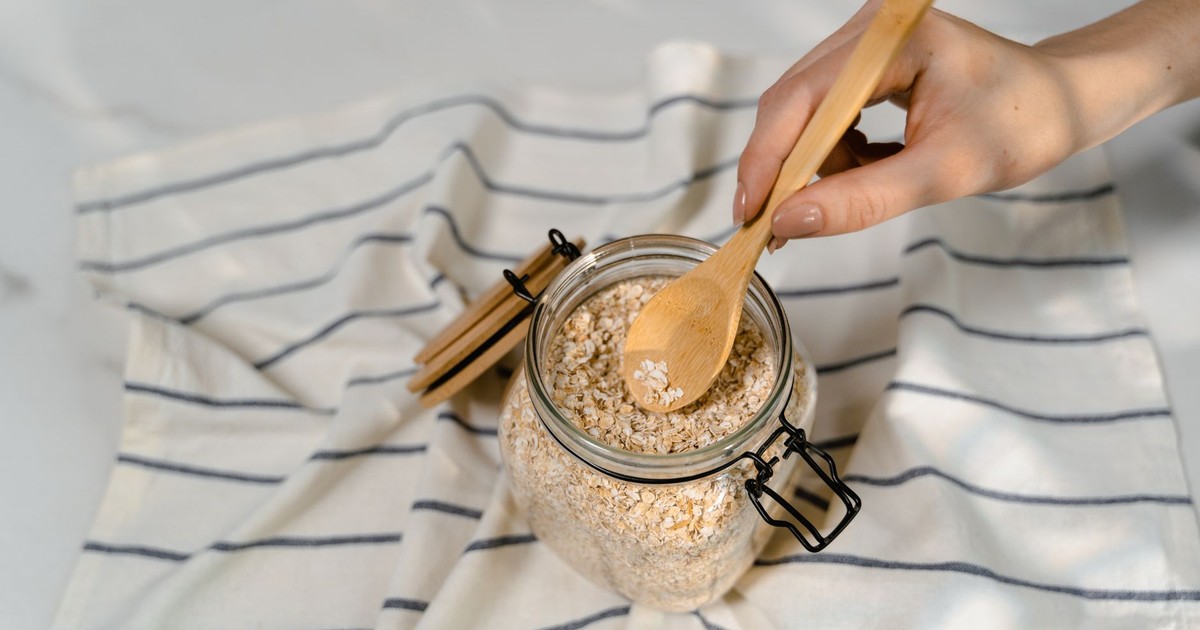 Close-up of a hand using a wooden spoon to scoop uncooked oatmeal from a glass jar on a striped cloth.