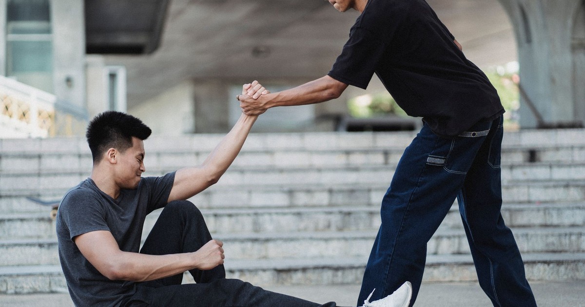 Side view Asian man in street wear holding friends hand and helping to get up from ground on sunny street