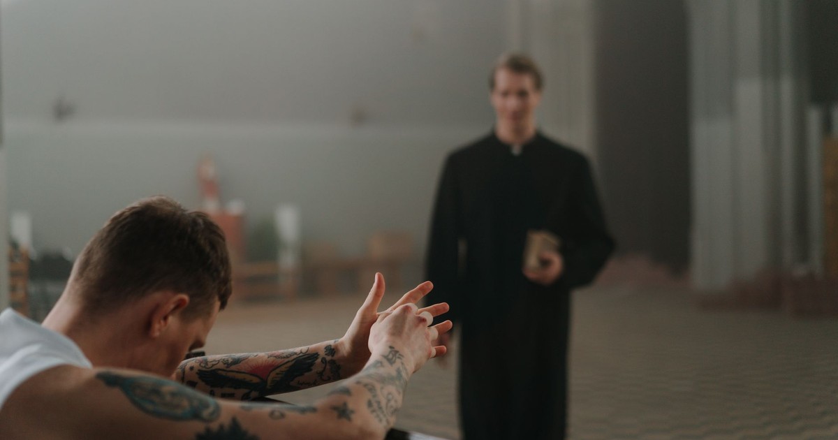A tattooed man prays in a church as a priest observes, capturing faith and reflection indoors.
