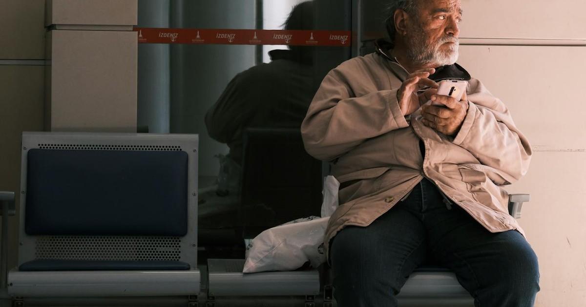 Elderly man holding a coffee cup, sitting alone on a bench indoors.