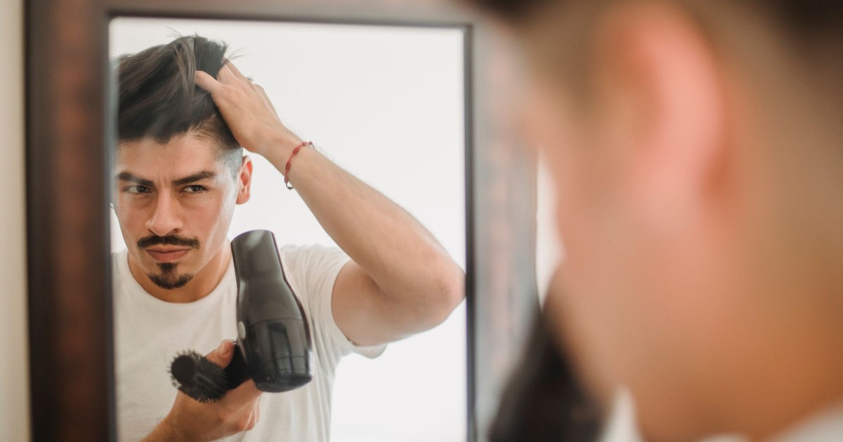Adult man styles his hair using a hairdryer while looking at his reflection, capturing a modern grooming routine.