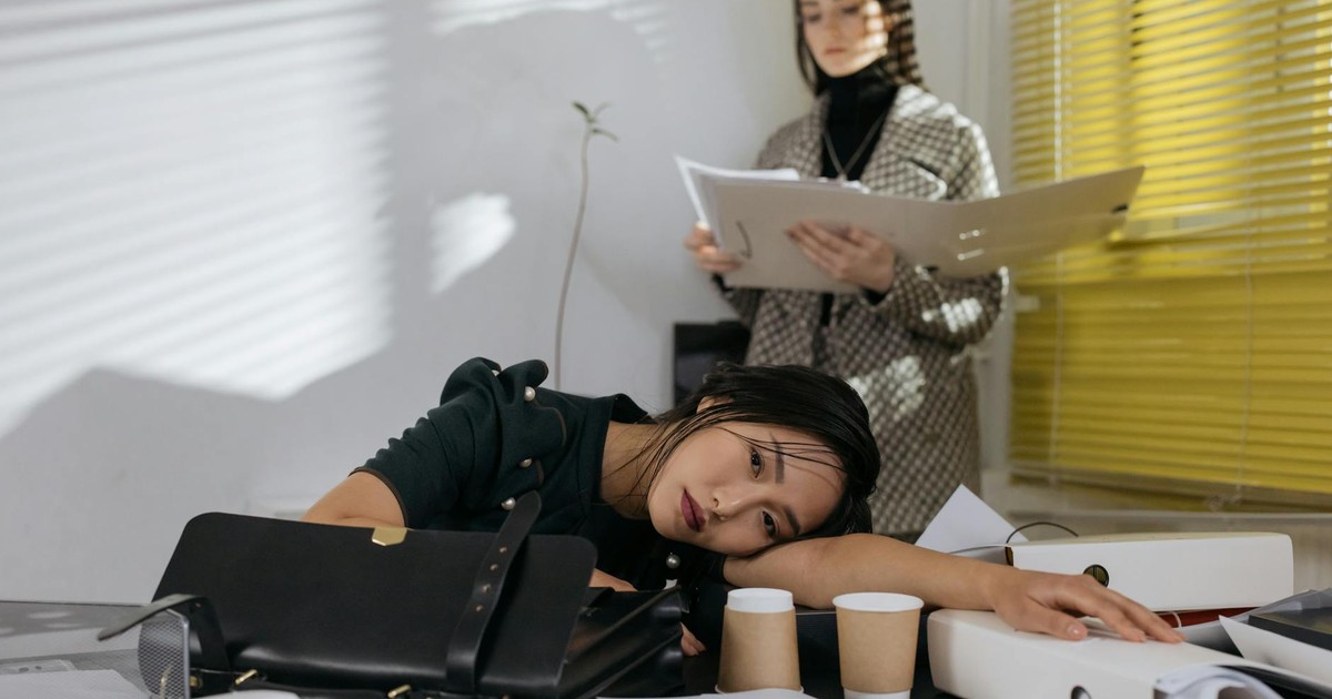 Exhausted employee resting on desk surrounded by documents, colleague in background.