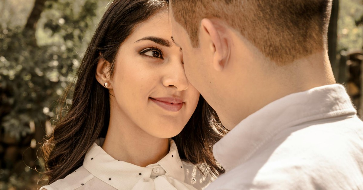 A young couple stands face to face, sharing a loving moment under a tree in daylight.