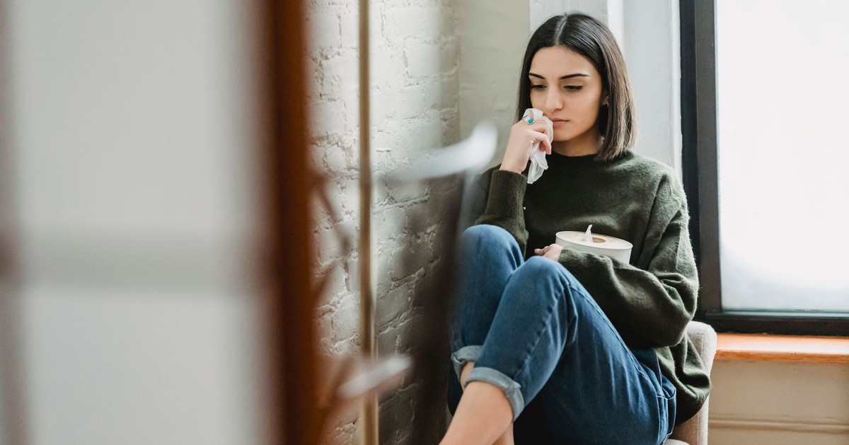 Upset young ethnic lady with dark hair in casual clothes sitting on comfortable armchair with tissues box in hand and looking down thoughtfully