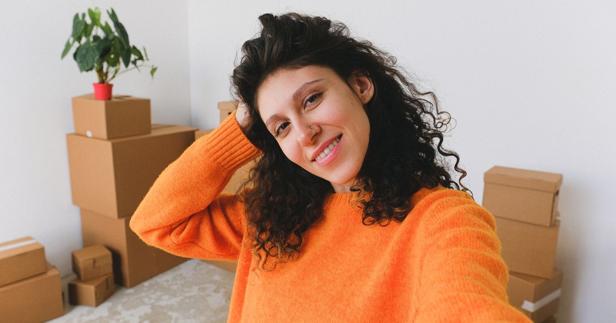 Cheerful young female smiling and touching curly hair while taking selfie against pile of cardboard boxes after relocation in new apartment