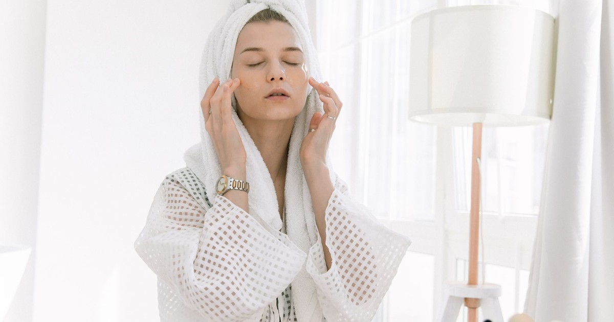 Adult woman in bathrobe doing skincare routine with head towel and under-eye patches indoors.