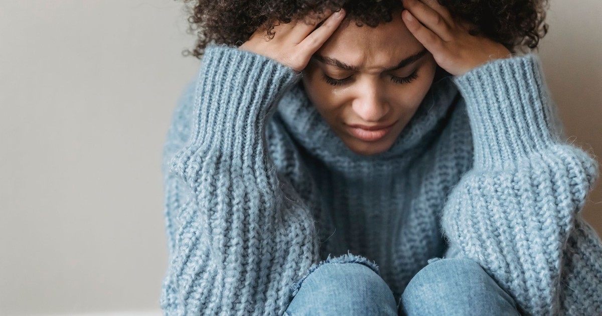 Frowning African American female with short curly hair in casual clothes sitting on floor and touching head against light wall