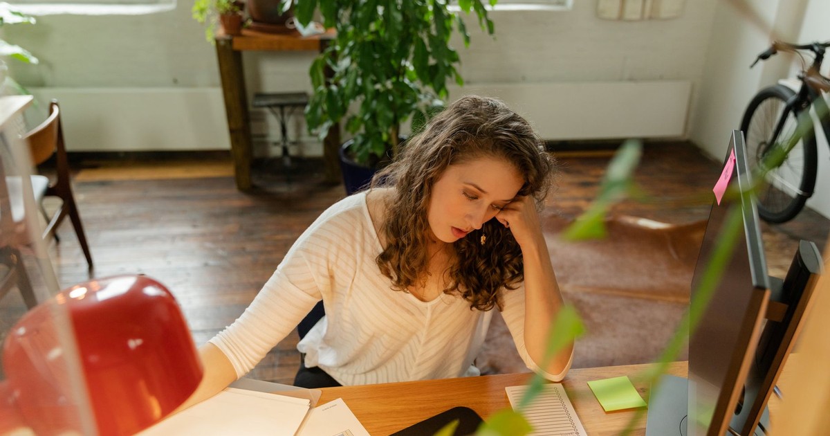 A woman sits at a wooden desk, appearing tired amid office work with sticky notes and a monitor.