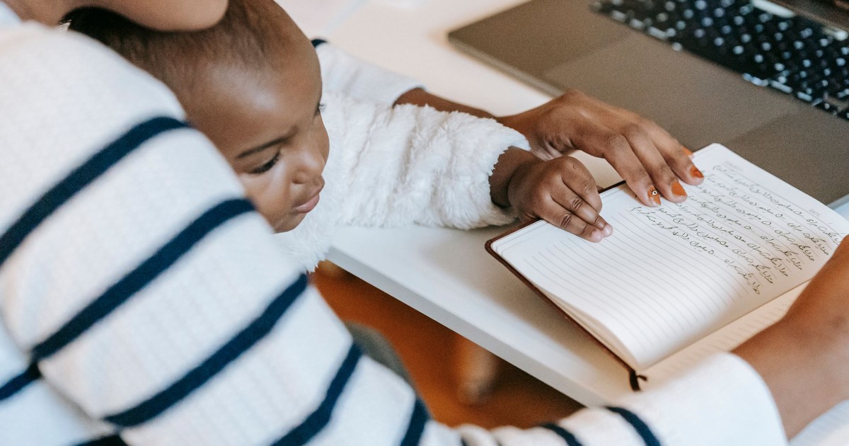 Ethnic baby with mother wearing casual outfit while sitting at table with opened notebook near laptop in bright room