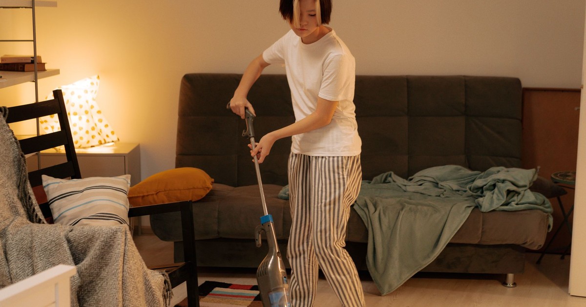 Asian woman cleaning a messy living room at home with a vacuum cleaner, creating a tidy space.