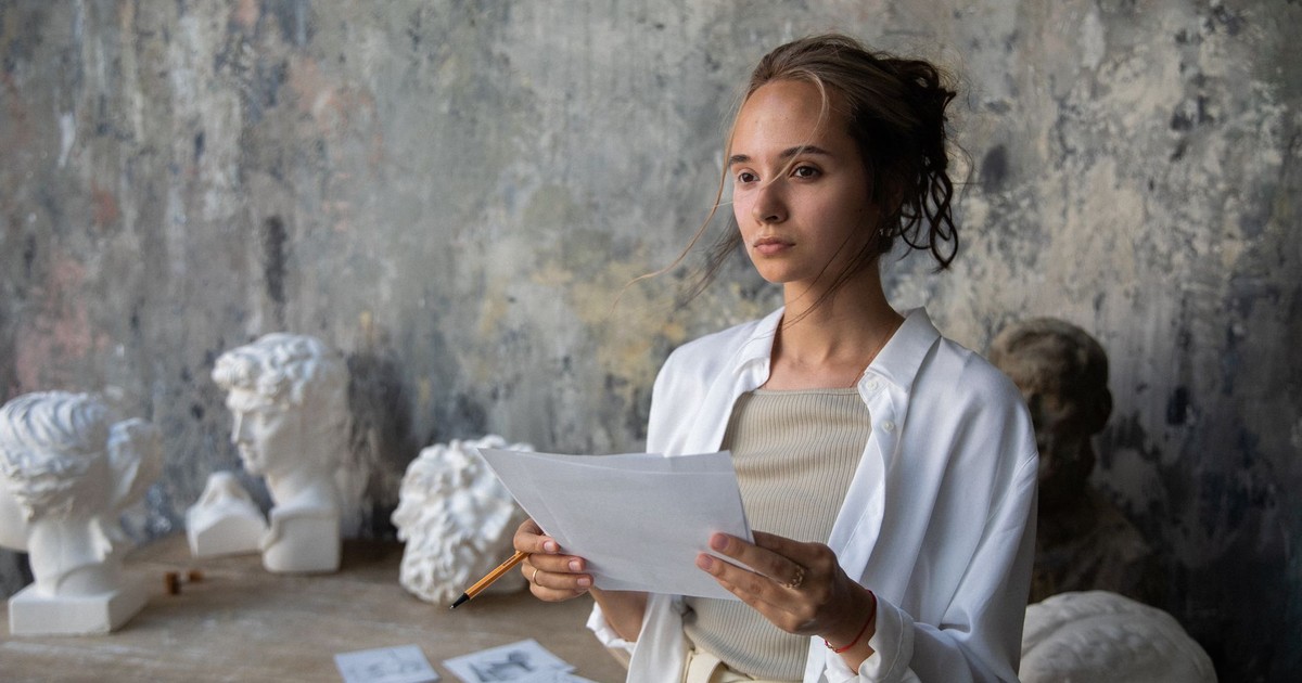 Woman in art studio holding paper, surrounded by classical sculptures.