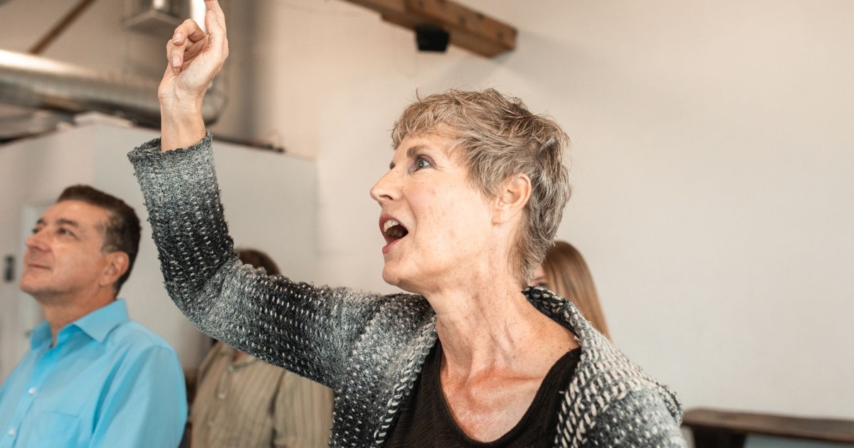 Senior caucasian woman attentively pointing during indoor seminar with diverse audience.