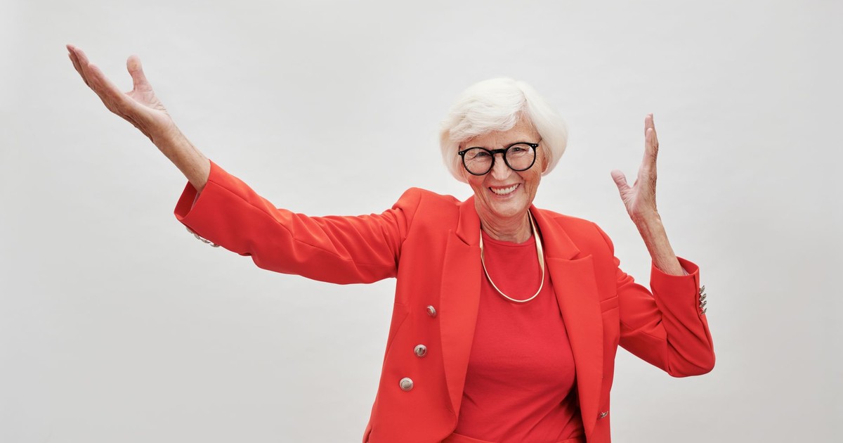 A joyful senior woman with white hair and eyeglasses in a red suit poses against a simple background.