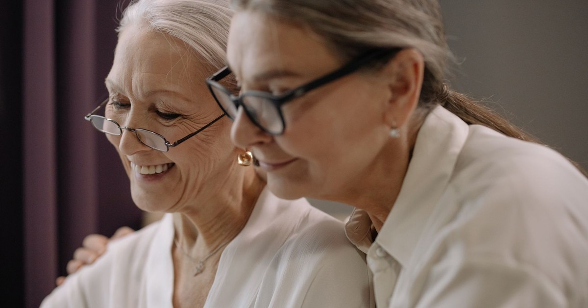 Two elderly women wearing glasses share a joyful moment indoors.