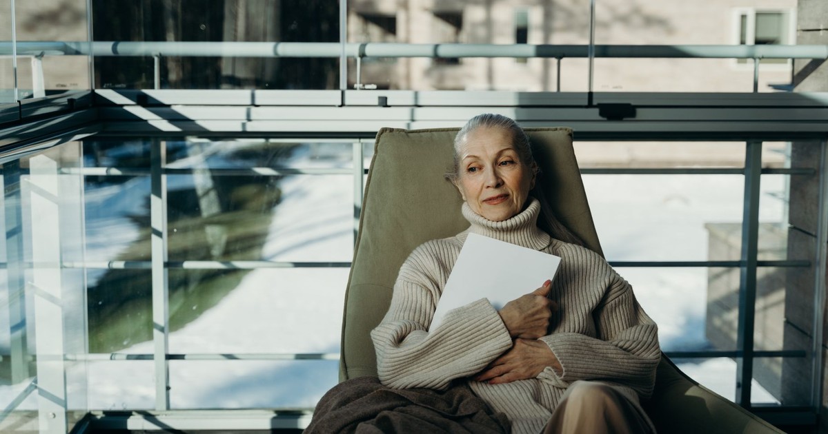 Elderly woman in cozy sweater relaxes indoors with a book in a serene setting.