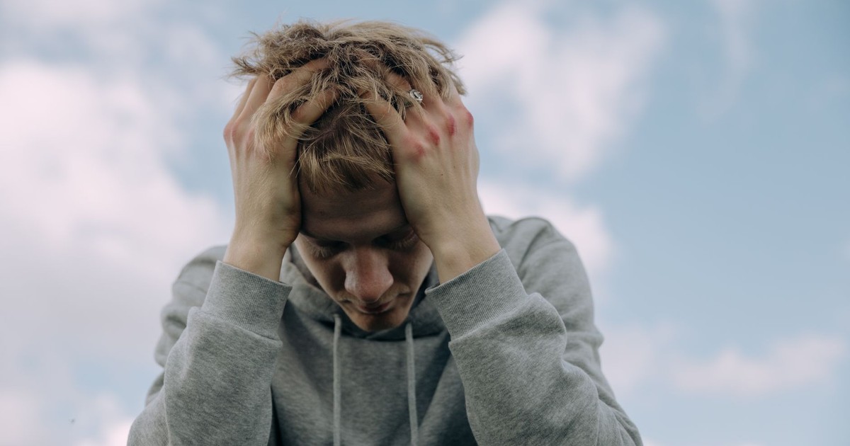 Young man in gray hoodie holds head in frustration, set against cloudy sky.