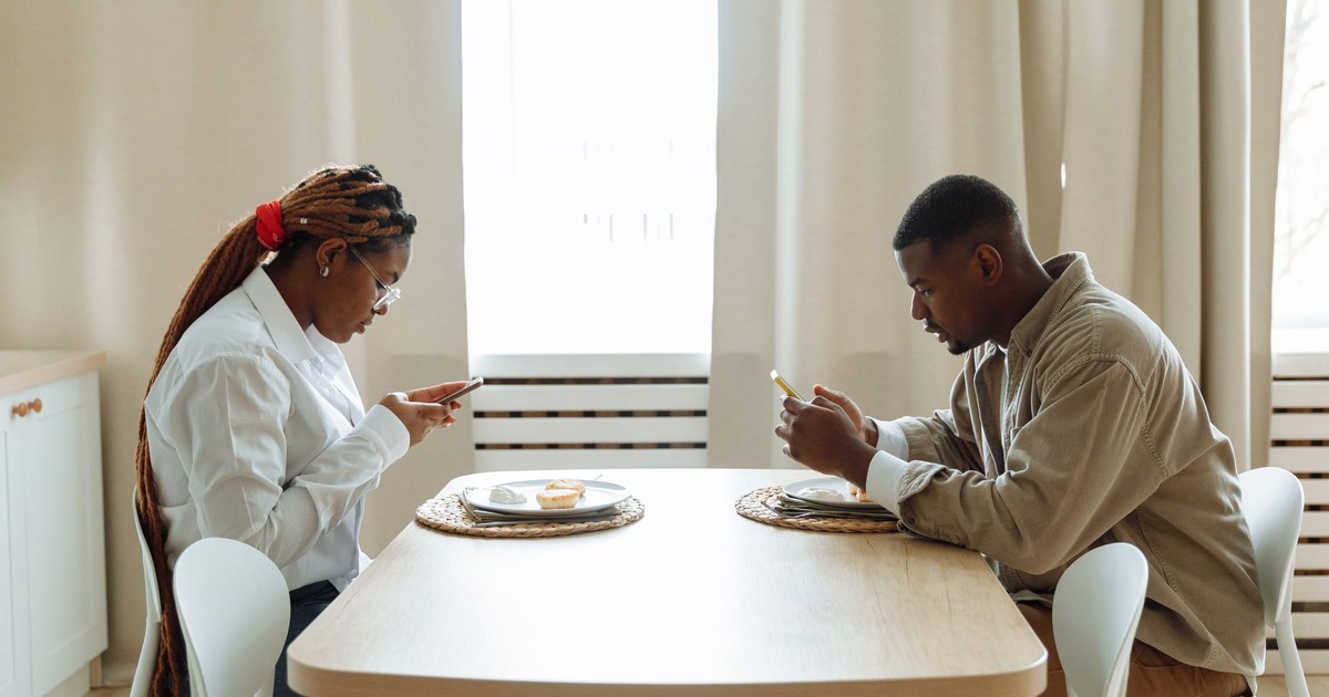 Two adults sitting at a dining table, focused on their smartphones while having breakfast indoors.