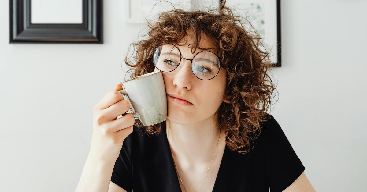 A thoughtful woman with curly hair and glasses holding a coffee mug in a modern room.