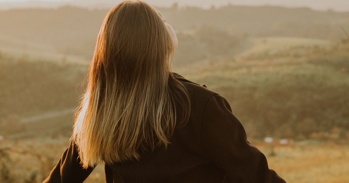 Back view of a woman with long hair standing in a scenic hillside at sunset, embracing nature.