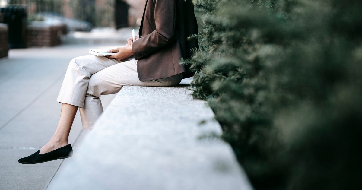 Side view of crop unrecognizable young ethnic female employee in classy outfit sitting on stone border in city park and taking notes in planner