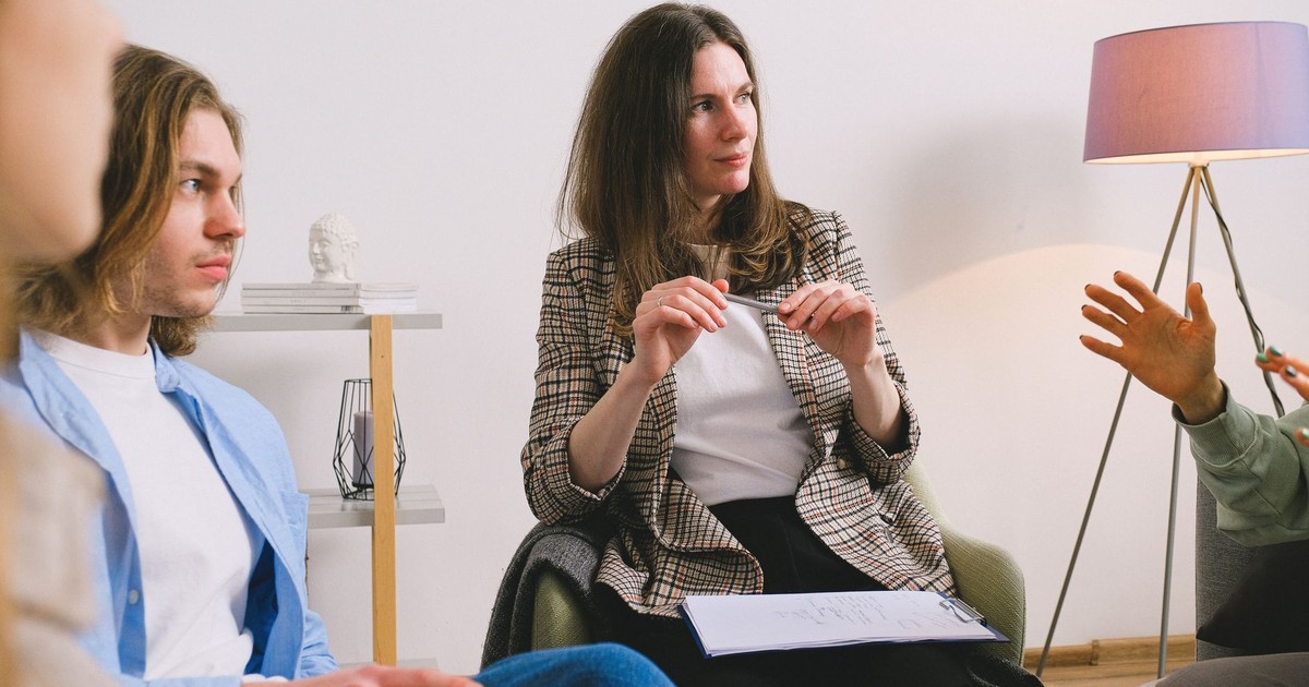 Pensive female sitting with clipboard on armchair and listening colleagues in light room in daytime